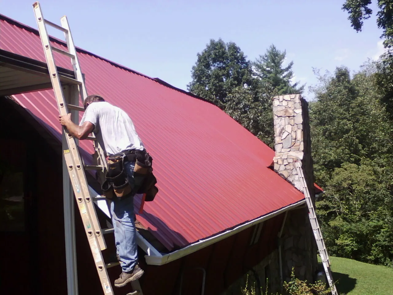 Roofer on a ladder beside a red metal roof and stone chimney — Soffit & Fascia Repair by Roofing Brothers in Patricksburg
